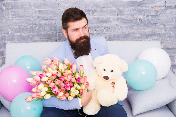 Romantic man with flowers and teddy bear sit on couch with air balloons waiting girlfriend. Romantic gift. Macho ready romantic date. Man wear tuxedo bow tie hold bouquet. She deserve all best