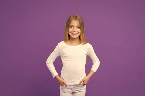 白いジャンパーとジーンズを着て子供、若者のファッションのコンセプトに。長い輝くブロンドの髪を持つ少女。紫色の背景に分離された笑顔と素敵な子供。かわいい顔とヘーゼル色の目の女子高生 — ストック写真