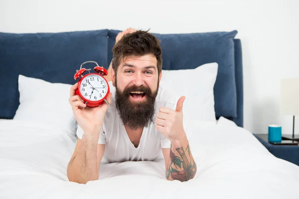 Enjoying leisure time at home. happy bearded man hipster with alarm clock. brutal sleepy man in bedroom. male with beard in pajama on bed. asleep and awake. energy and tiredness. time management