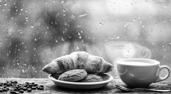 Enjoying coffee on rainy day. Coffee time on rainy day. Fresh brewed coffee in white cup or mug on windowsill. Wet glass window and cup of hot caffeine beverage. Coffee drink with croissant dessert — Stock Photo, Image