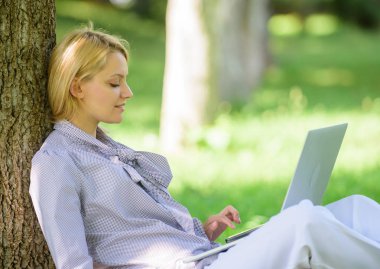 Kız parkta dizüstü bilgisayar ile çim üzerinde oturup çalışır. Doğal çevre ofisi. Açık havada çalışmanın faydaları. Eğitim teknolojisi ve internet konsepti. Dizüstü bilgisayar işi açık havada kadın ağaç gövdesi yalın