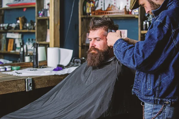 Conceito de estilo de vida Hipster. Um cliente Hipster a cortar o cabelo. Barbeiro com cortador de cabelo trabalhar no penteado para hipster, barbearia fundo. Barbeiro com clipper aparar o cabelo no templo do cliente barbudo — Fotografia de Stock