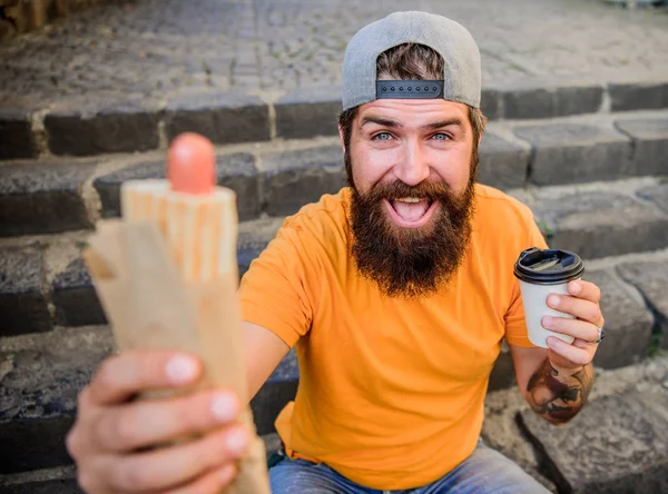 Um lanche de homem faminto. Um tipo a comer cachorros quentes. Homem barbudo mordida saborosa salsicha e beber copo de papel. Comida de rua tão boa. Nutrição de estilo de vida urbano. Comida de plástico. Despreocupado hipster comer junk food while sit on stairs — Fotografia de Stock