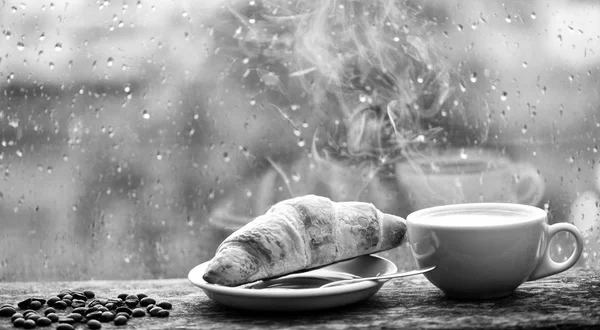 Koffie drinken met croissant dessert. U geniet van koffie op regenachtige dag. Koffie tijd op regenachtige dag. Vers gebrouwen koffie in witte kopje of mok op de vensterbank. Natte glazen raam en kopje warme cafeïne drank — Stockfoto