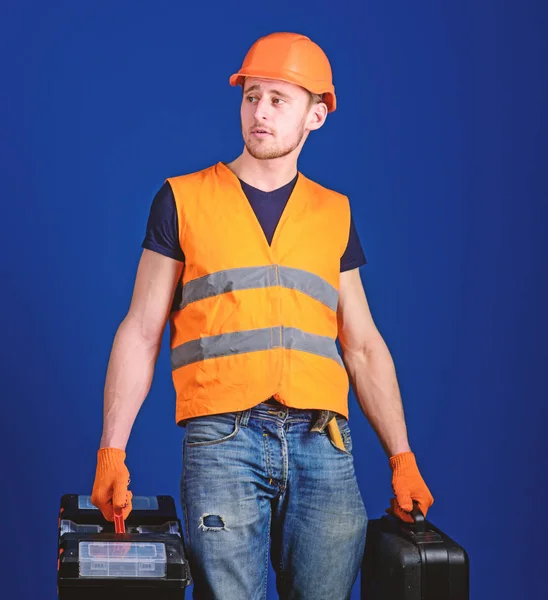 Concetto riparatore attrezzato. Uomo in casco, cappello rigido porta attrezzi e valigia con attrezzi, sfondo blu. Operaio, tuttofare, riparatore, costruttore su viso calmo porta borse con strumenti professionali — Foto Stock