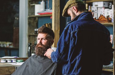 Sakallı hippi şık berber dükkanı olarak profesyonel kuaför tarafından hizmet veren cape kaplı. Damat kavramı. Sakal hipster istemcisinin damat ile meşgul berber, arka plan üzerinde güzellik malzemeleri
