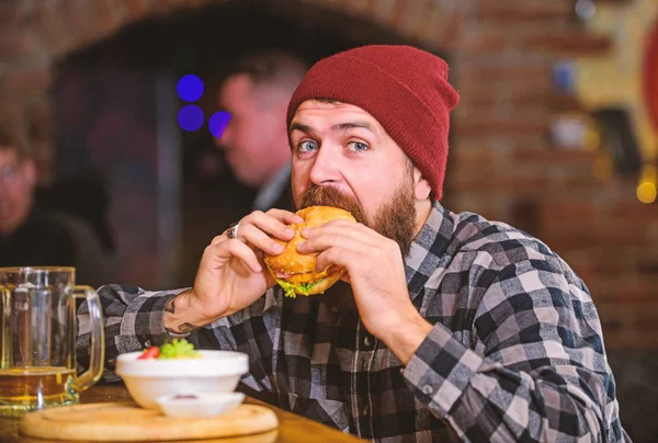 Brutal hipster bearded man sit at bar counter. High calorie food. Cheat meal. Delicious burger concept. Enjoy taste of fresh burger. Hipster hungry man eat burger. Man with beard eat burger menu