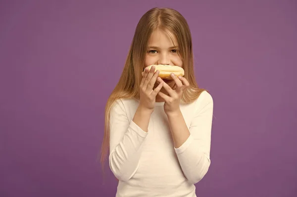 Comiendo bocadillos. El niño come rosquilla sobre fondo violeta. Niña con anillo esmaltado donut sobre fondo púrpura. Un chico con comida chatarra. Comida para merienda y postre. Infancia y cuidado de niños, espacio para copiar — Foto de Stock