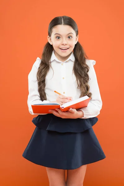 Diario de la escuela para tomar notas. sorprendió a una niña pequeña con uniforme escolar. obtener información libro de formularios. colegiala escribir notas sobre fondo naranja. de vuelta a la escuela. reportero escolar. carrera de reportero —  Fotos de Stock