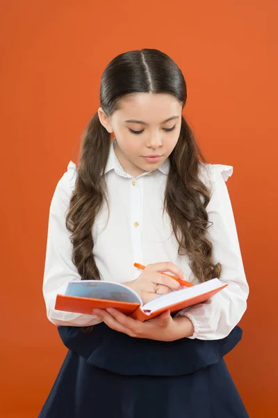 Schülerin schreibt Notizen auf orangefarbenem Hintergrund. Lesestunde. Holen Sie sich Informationen aus Buch. zurück zur Schule. Kleines Kind konzentrierte sich auf die Arbeit. kleines Mädchen in Schuluniform. Schreiben im Arbeitsbuch — Stockfoto