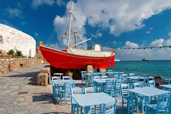 Ship and tavern furniture on quay in Mykonos, Greece. Red boat and blue tables on sea beach. Beach restaurant with sea view. Summer vacation on mediterranean island. Travelling for romance