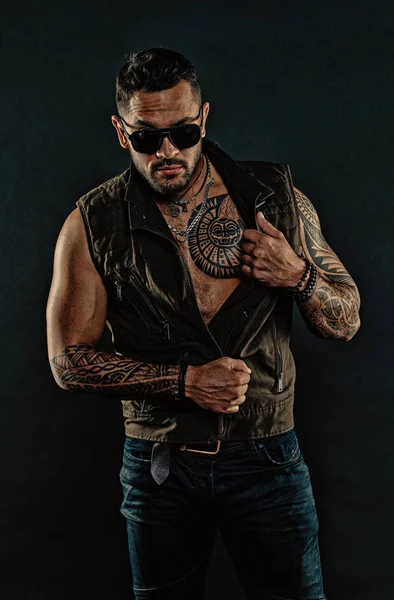 Fashion macho with tattoo on chest and arms. Fashion style and trend. Tattoo model with beard on unshaven face. Tattooed man with biceps and triceps. Bearded man in trendy sunglasses, vintage filter