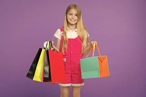 Gelukkig kind met boodschappentassen op violette achtergrond. Meisje glimlach met papieren zakken. Kid shopper in mode jumpsuit. Vakantie voorbereiding en viering. Na een dag winkelen. verkoop en zwarte vrijdag — Stockfoto