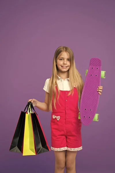 스케이트 보드와 보라색 배경에 쇼핑백 행복 소녀. 종이 가방 및 스케이트 보드 아이 미소 작은. 후 일 쇼핑. 판매와 블랙 금요일입니다. 스포츠 및 활성 게임 — 스톡 사진