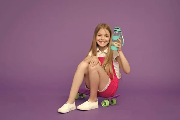 Staying hydrated. Girl happy face holds with water bottle while sits skateboard, violet background. Kid girl keeps staying hydrated. Body hydration concept. Girl cares about health and water balance
