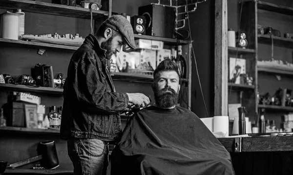 Grooming concept. Hipster with beard covered with cape serving by professional barber in stylish barbershop. Barber busy with grooming beard of hipster client, beauty supplies on background — Stock Photo, Image