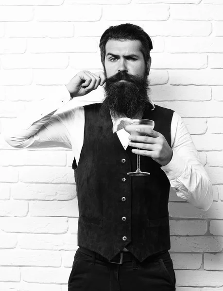 Handsome bearded man with long beard and mustache has stylish hair on serious face holding glass of alcoholic beverage in vintage suede leather waistcoat on white brick wall studio background — Stock Photo, Image