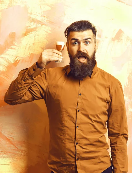 Bearded man, long beard. Brutal caucasian surprised hipster with moustache in brown shirt holding alcoholic red shot on colorful texture backgroun