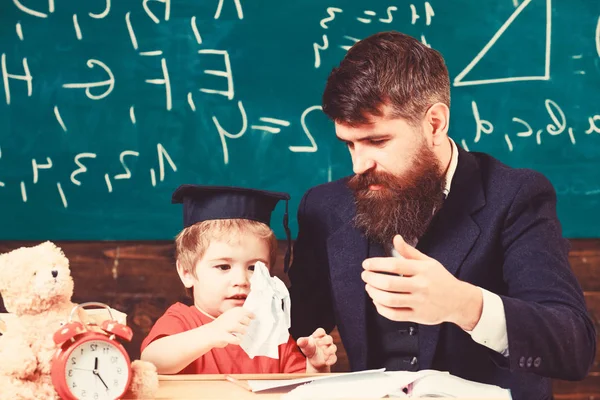 Leraar nieuw materiaal uit te leggen aan kleine leerling. Schattige jongen in mortarboard spelen met papier. Knappe jongen en schattige jongen voor de groene bestuur — Stockfoto