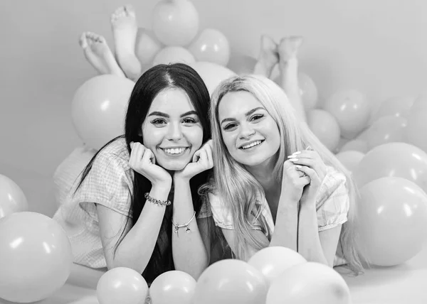 Concepto de chismes. Rubia y morena en caras sonrientes se divierten en la fiesta del dormitorio doméstico. Las niñas yacían sobre el vientre cerca de globos, fondo rosa. Hermanas o mejores amigos en pijama en fiesta de pijamas de niña —  Fotos de Stock