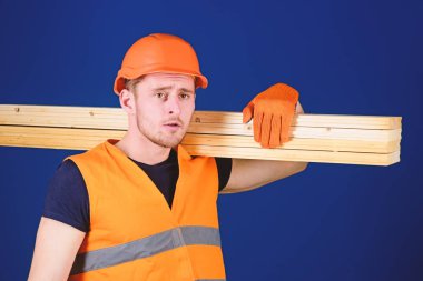 Adam kask, kask ve koruyucu eldiven tutan ahşap kiriş, mavi arka plan. Ahşap malzemeler kavramı. Marangoz, woodworker, düşünceli yüz taşıdığı ahşap kiriş omuz üzerinde güçlü Oluşturucu