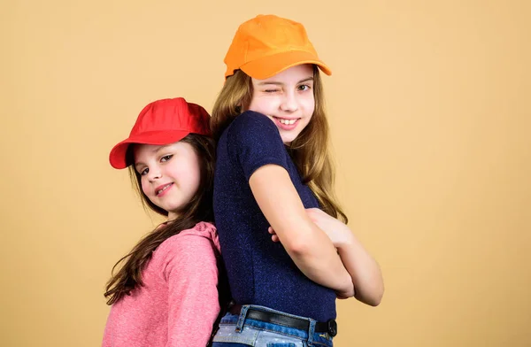 Cool girls. Sisters stand back to back beige background. Little cute girls wearing bright baseball caps. Modern fashion. Hats and caps. Stylish accessory. Kids fashion. Feeling confident wearing caps