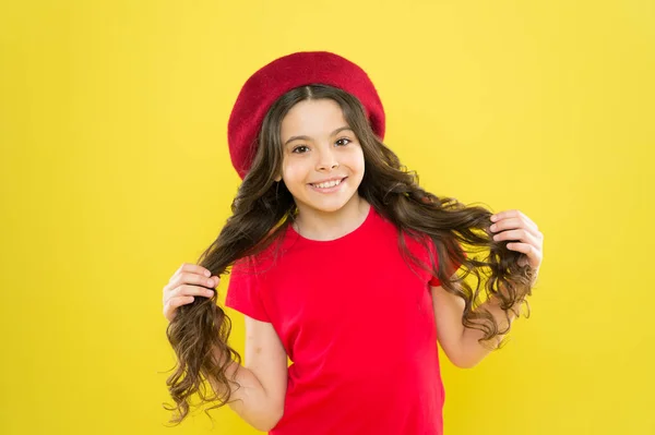 Sorte e beleza. Criança feliz rosto bonito adorável cabelo encaracolado fundo amarelo. Dicas de beleza para cabelos arrumados. Criança sorridente. Kid menina longo cabelo brilhante saudável usar chapéu vermelho. Menina com cabelo comprido — Fotografia de Stock