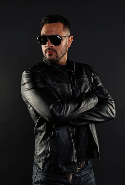 Confident man stand with arms folded. Bearded man in trendy sunglasses. Fashion model in leather jacket and jeans. Fashion and style. Confidence with sexuality and charisma