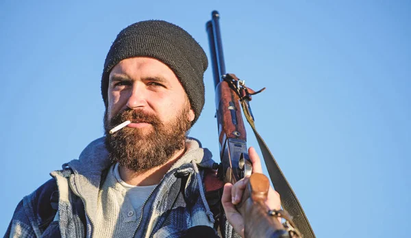 Hunting masculine hobby concept. Man brutal bearded guy gamekeeper blue sky background. Brutality and masculinity. Hunter with rifle gun close up. Guy bearded hunter spend leisure hunting and smoking