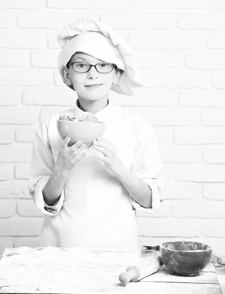Kleiner Junge netter Koch Koch in weißer Uniform und Hut auf gefärbtem Gesicht Mehl mit Gläsern, die neben Tisch mit Nudelholz stehen und halten türkisfarbene Schüssel mit Schokoladenkeksen auf Backsteinwand Hintergrund — Stockfoto