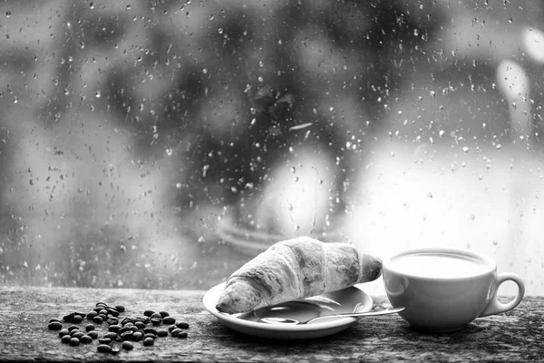 Natte glazen raam en kopje warme cafeïne drank. Koffie drinken met croissant dessert. U geniet van koffie op regenachtige dag. Koffie tijd op regenachtige dag. Vers gebrouwen koffie in witte kopje of mok op vensterbank — Stockfoto