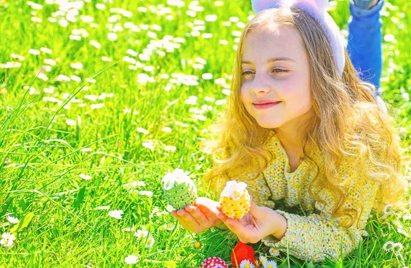 Child with cute bunny ears lying at meadow. Cute child lay on grass with egg in hands. Little girl hunting for Easter egg in spring garden on Easter day, traditional celebration. Easter feast concept