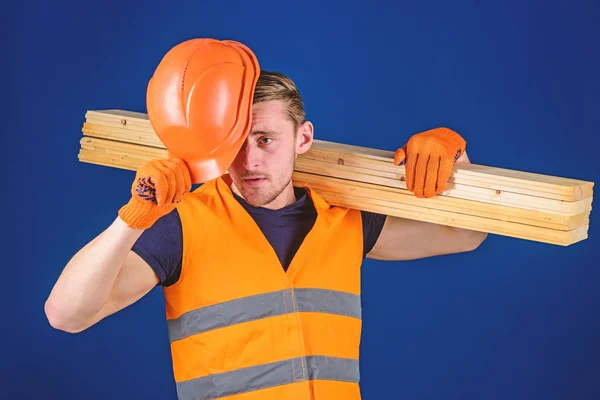 Protective equipment concept. Carpenter, woodworker, labourer, builder carries wooden beam on shoulder. Man in protective gloves holds visor of helmet, corrects hard hat on head, blue background — Stock Photo, Image