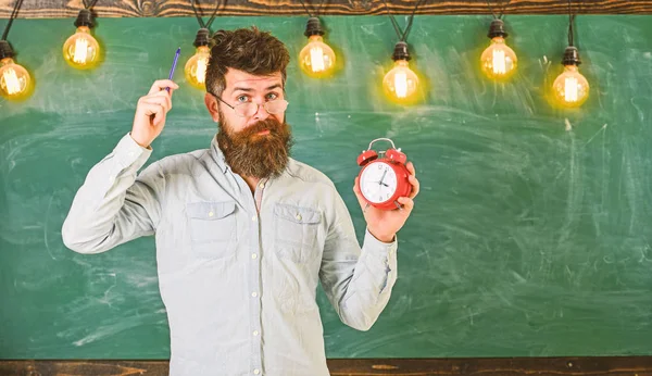 Homem com barba e bigode na cara esquecida comichão na cabeça. O hipster barbudo segura relógio, quadro no fundo, espaço de cópia. Conceito de disciplina. Professor em óculos detém despertador e caneta — Fotografia de Stock