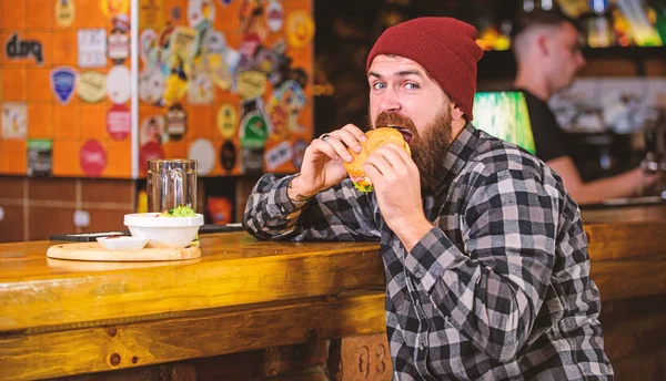 Delicious burger concept. Enjoy taste of fresh burger. Hipster hungry man eat burger. Man with beard eat burger menu. Brutal hipster bearded man sit at bar counter. Cheat meal. High calorie food