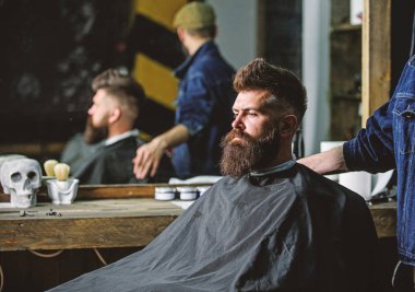 Siyah pelerin ile kaplı sakallı salonların sandalye, ayna arka plan oturur. Hipster berber sakal müşterilerinden olan adam. Berber kavramı. Sakallı hippi Kuaför ve saç kesimi için bekler