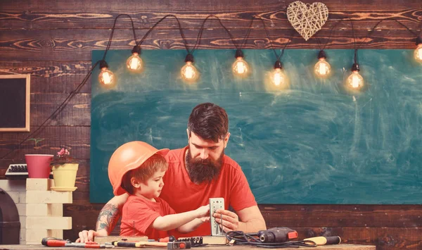 Fathers assistant concept. Father with beard teaching little son to use tools in classroom, chalkboard on background. Boy, child busy in protective helmet makes by hand, repairing with dad