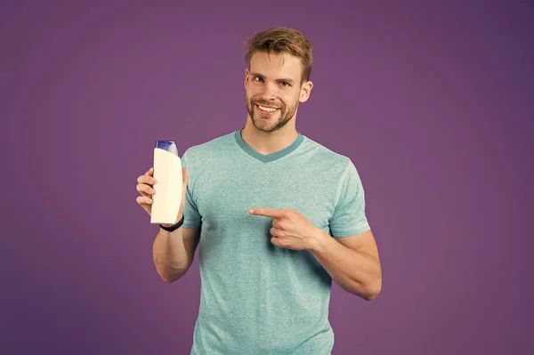 Cuidado del cabello y piel sana sin caspa. Hombre feliz cara sonriente apuntando a fondo violeta botella de plástico. Chico con cerdas recomiendan champú botella. Hombre disfrutar de frescura lavado de champú para el cabello — Foto de Stock