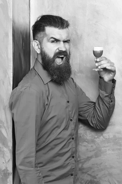 Bearded man, long beard. Brutal caucasian happy hipster with moustache in brown shirt holding alcoholic red shot on golden texture backgroun