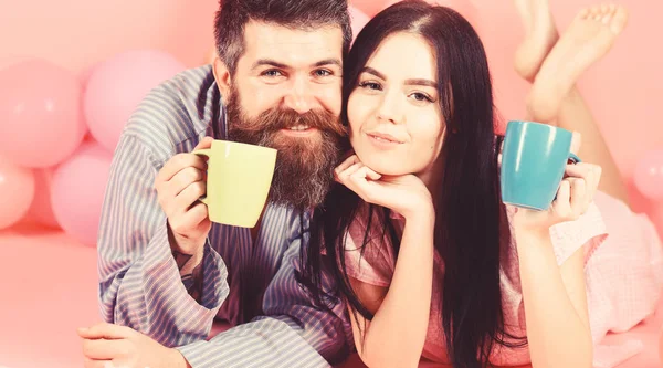 Hombre y mujer en rostros sonrientes yacían, fondo rosa. Pareja relajarse en la mañana con café. Pareja enamorada bebe café en la cama. Concepto de tradición familiar. Hombre y mujer en ropas domésticas, pijamas — Foto de Stock