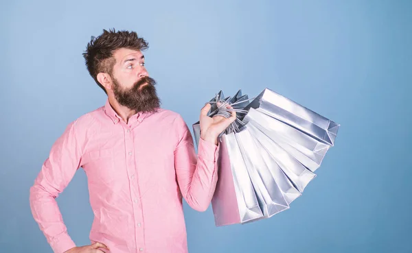 Sale and discount concept. Hipster on serious face carries shopping bags. Guy shopping on sales season with discounts. Man with beard and mustache works as shop assistant, light blue background