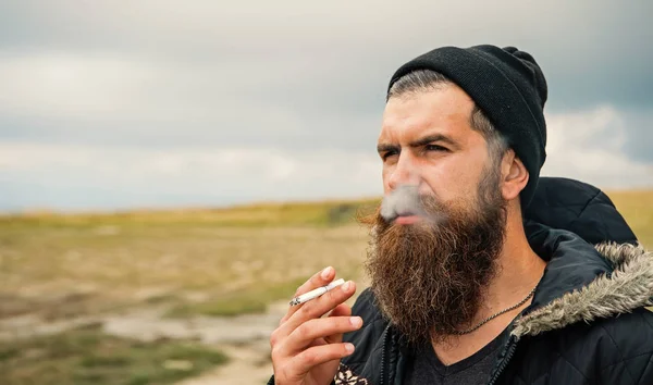 Hombre barbudo humo cigarrillo en sombrero al aire libre sobre fondo natural — Foto de Stock