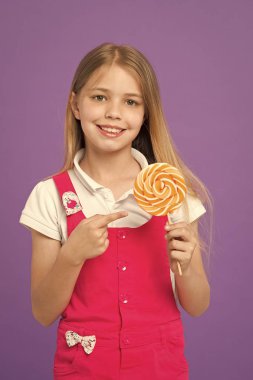 Sevimli neşeli kız dev şeker tutarak. Menekşe arka plan üzerinde izole uzun sarı saçlı çocuk. Renkli lolipop, tatlı çocukluk kavramı işaret eden çocuk. Pembe tulum Madem Hadleyberg'deki tatlılar seviyor.