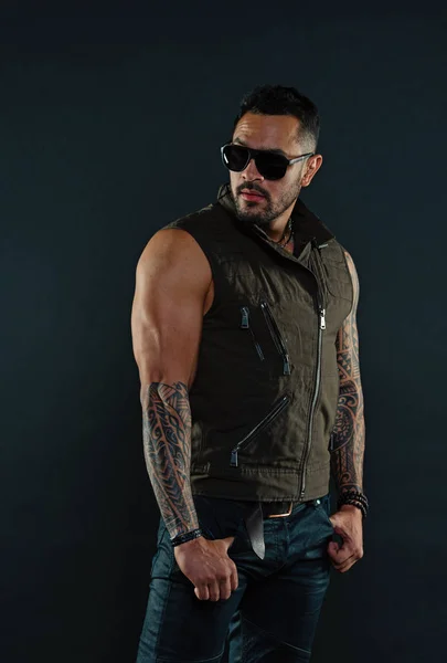 Bearded man with tattoo on strong arms. Tattoo model with beard on unshaven face. Tattooed man with biceps and triceps. Fashion macho in trendy sunglasses. Fashion style and trend