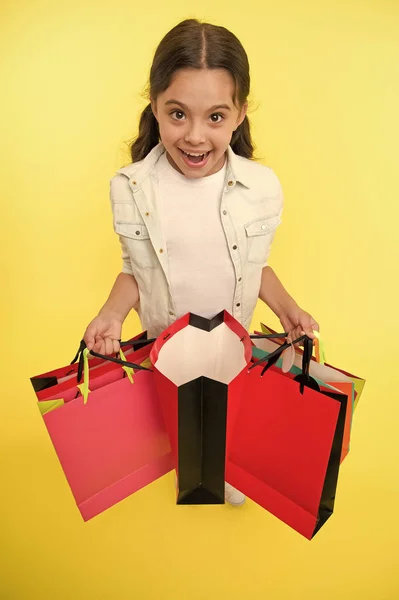 Nézd meg a nyereséges vásárlásait. A lány sárga hátteret hord magánál. A lány szeret vásárolni. Gyermek aranyos boltos egy csomó bevásárló táskával fekete péntek teljes kiárusítás. Izgalmas vásárlás — Stock Fotó