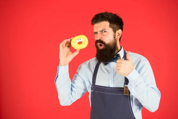 Hipster padeiro barbudo segurar donut vitrificado no fundo vermelho. Conceito de café e padaria. Doce donut do padeiro. Homem padeiro barbudo em avental segurar sobremesa bonito. Negócios privados de padaria. Experimenta isto. — Fotografia de Stock