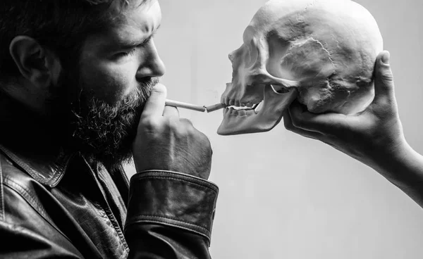 Man smoking cigarette near human skull symbol of death. Harmful habits. Destroy your health. Smoking is harmful. Habit to smoke tobacco bring harm to your body. Smoking cause health damage and death