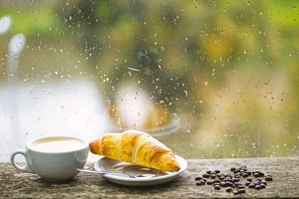 Wet glass window and cup of hot caffeine beverage. Coffee drink with croissant dessert. Enjoying coffee on rainy day. Coffee time on rainy day. Fresh brewed coffee in white cup or mug on windowsill