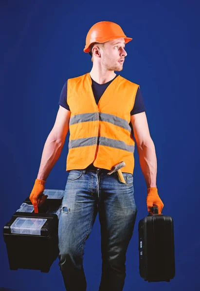 Conceito de reparador profissional. Homem de capacete, chapéu duro segura caixa de ferramentas e mala com ferramentas, fundo azul. Handyman, reparador na cara concentrada carrega sacos pesados com equipamento profissional — Fotografia de Stock