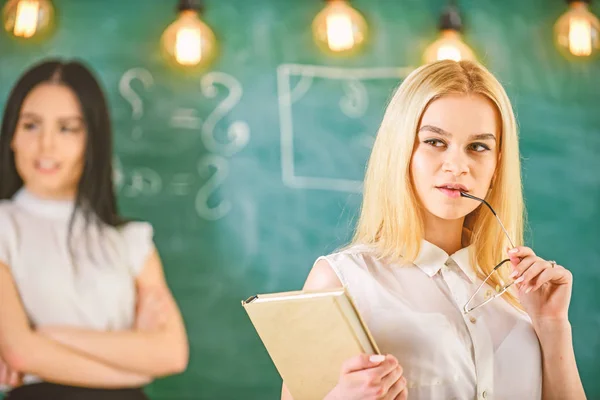 Chica celosa del éxito de compañero de clase en el aula, pizarra en el fondo. La mujer con libro y anteojos se ve soñadora mientras que la estudiante de fondo se ve celosa. Concepto de envidia y competencia —  Fotos de Stock